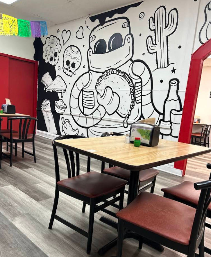 The interior mural at Tacos El Carnalito in Wilmington — a black and white artwork featuring a taco-holding astronaut, Day of the Dead skulls, and cactus